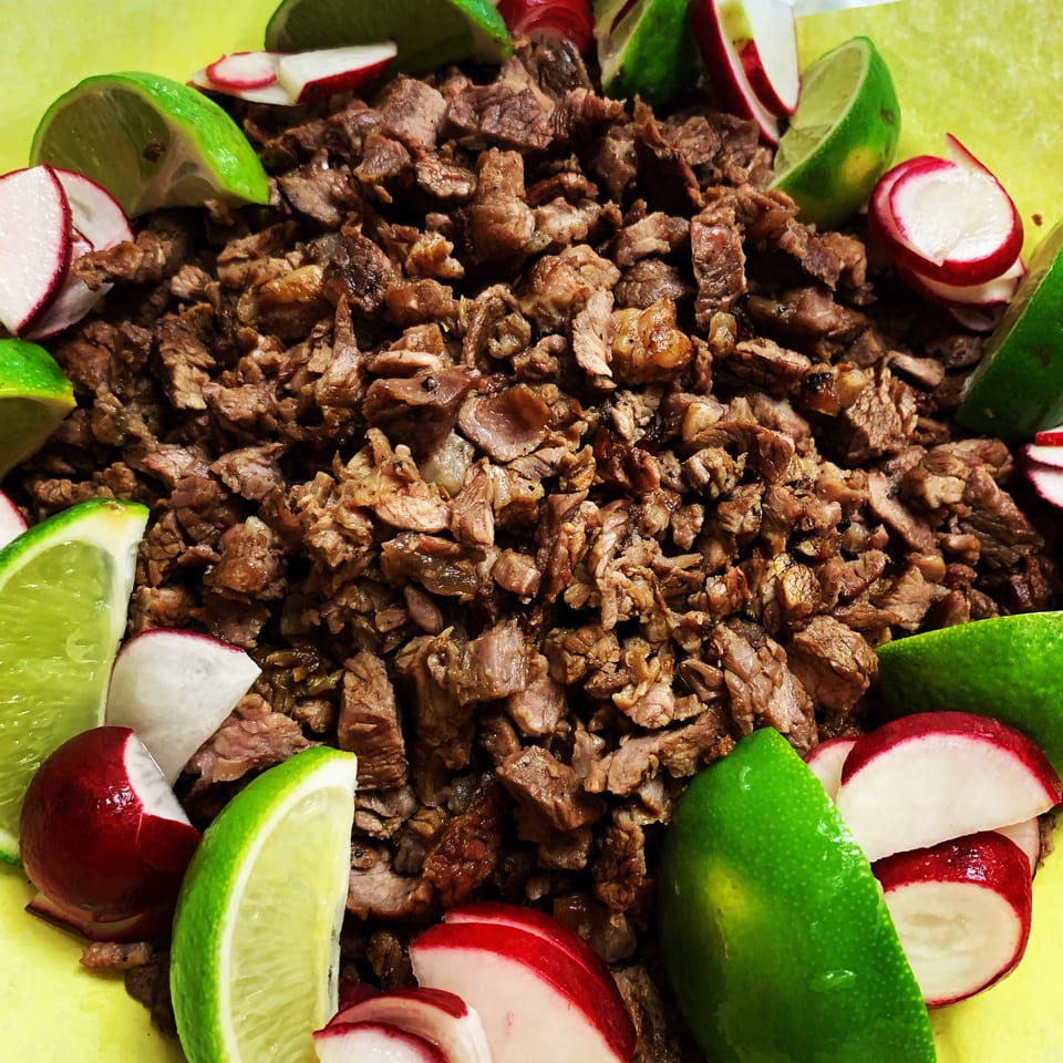 Close-up of diced carne asada, garnished with lime wedges and sliced radishes on a yellow surface.