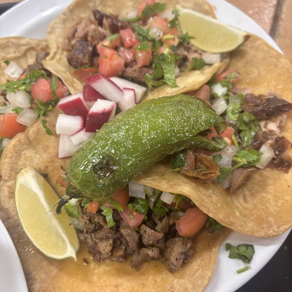 A plate of tacos with meat, pico de gallo, radishes, lime wedges, and a roasted green pepper.