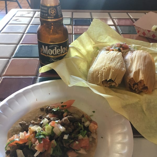 A Modelo Negra beer, a taco, and two tamales in corn husks on a tiled table.
