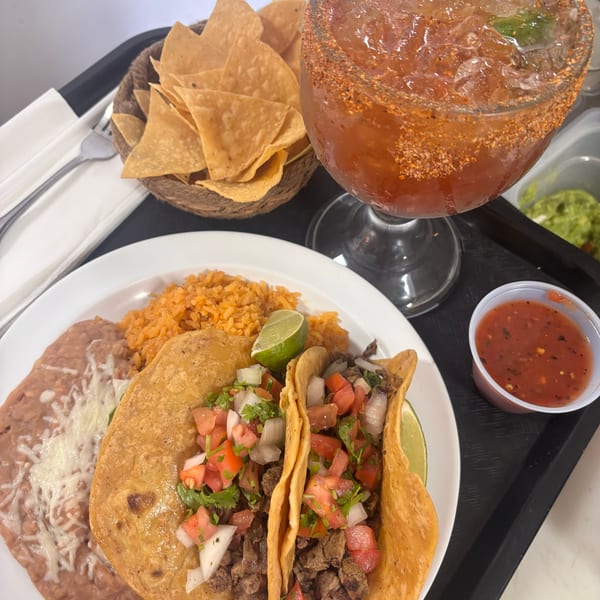 Two tacos with rice and beans, a basket of chips, and a large drink on a black tray.
