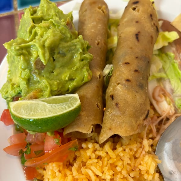 Two taquitos with guacamole, rice, pico de gallo, and a lime wedge on a white plate.