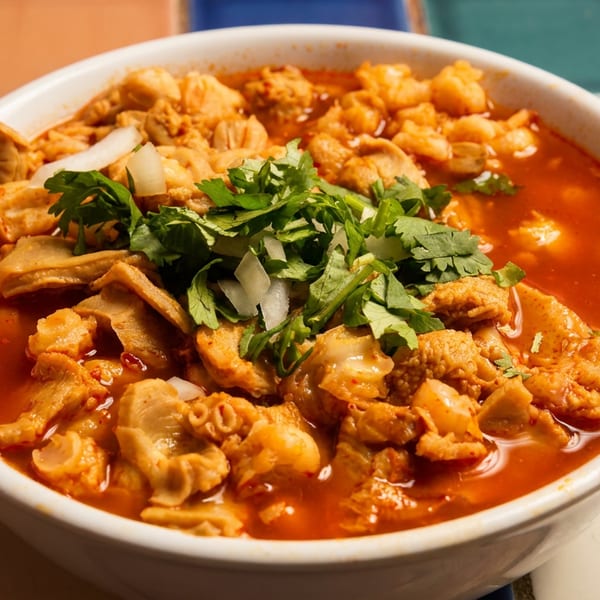 A white bowl of menudo soup with red broth, tripe, hominy, cilantro, and onions.