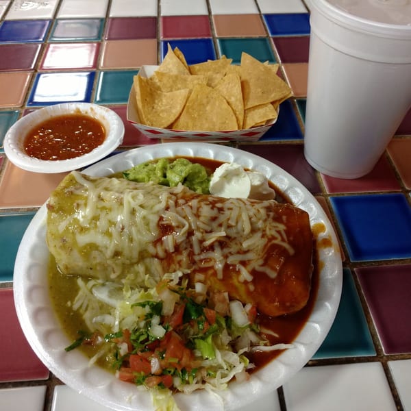 A large burrito covered in red and green sauce, with guacamole, sour cream, chips, and salsa.
