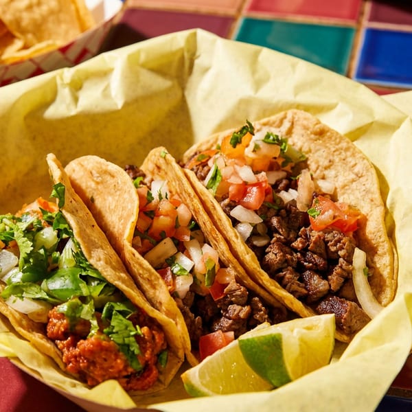 Three vibrant tacos with various fillings, lime, chips, and salsa on a colorful tiled table.
