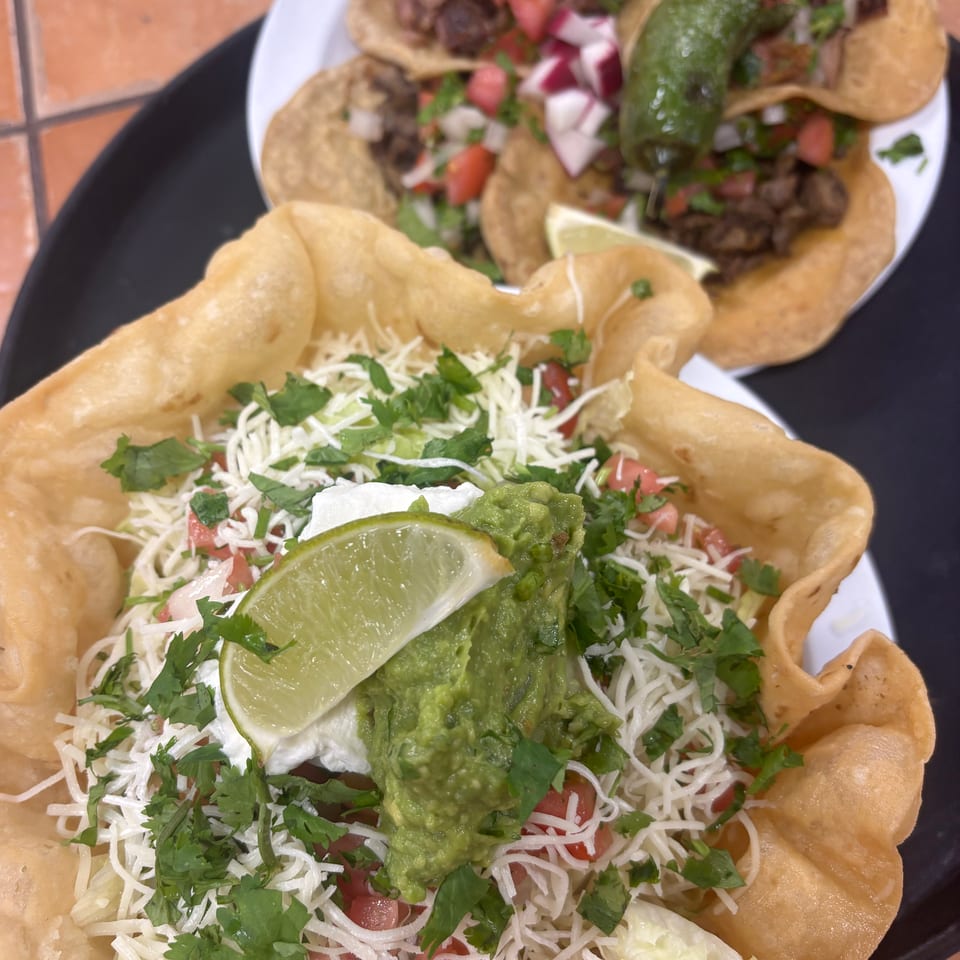 A taco salad in a fried tortilla bowl with guacamole, sour cream, and lime, next to street tacos.