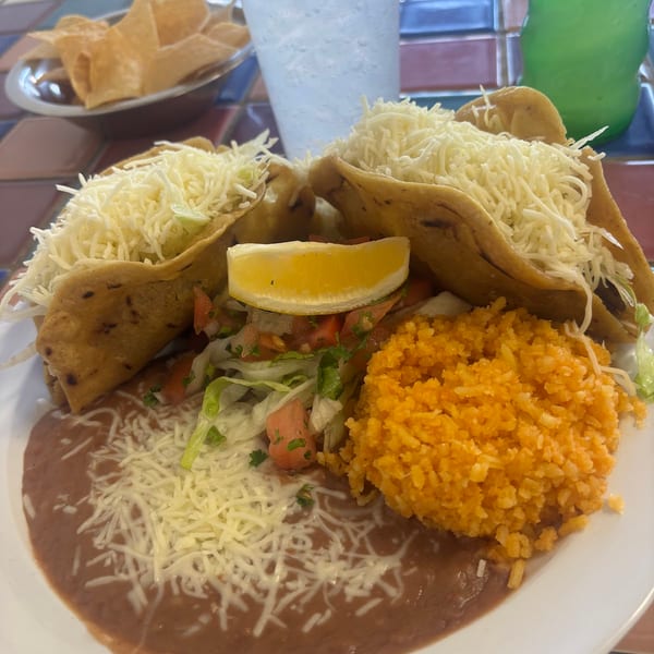 Three crispy tacos topped with shredded cheese, served with rice, refried beans, and a lemon wedge.