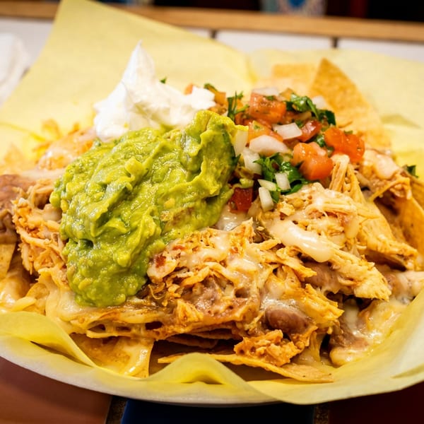 Chicken nachos with guacamole, sour cream, and pico de gallo in a yellow paper-lined basket.